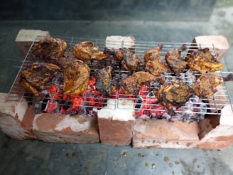 Grilled chicken pieces are arranged on a metal grate over glowing red charcoal, supported by bricks on either side. The chicken is seasoned with spices, giving it a charred, golden brown appearance.