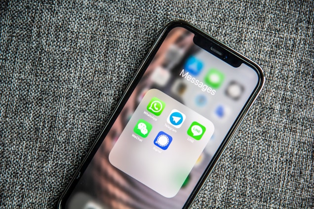 A smartphone resting on a textured fabric surface displays a folder labeled 'Messages' with multiple messaging app icons arranged within. The apps visible are WhatsApp, Telegram, LINE, WeChat, and Signal, each represented by their distinct logos.