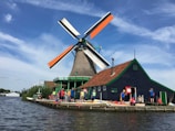 A traditional Dutch windmill with orange and white blades stands prominently beside a dark green house with a red roof. The scene includes several people dressed in bright clothing, likely tourists or visitors, along the bank of a wide, calm river. The sky above is blue with scattered wispy clouds.