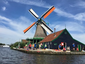 A traditional Dutch windmill with orange and white blades stands prominently beside a dark green house with a red roof. The scene includes several people dressed in bright clothing, likely tourists or visitors, along the bank of a wide, calm river. The sky above is blue with scattered wispy clouds.