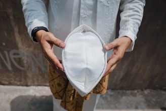 A person wearing a white traditional outfit is holding a white hat with both hands, positioned in the center of the image. The background is neutral with some faint writing on the wall.