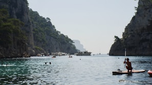A scenic waterway with steep rocky cliffs covered in greenery on both sides. The water is clear and blue, dotted with several people swimming. In the foreground, there is a person on a paddleboard holding a paddle. The background features a few boats anchored in the distance.