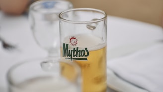 A clear glass filled with golden beer, branded with the logo 'Mythos,' is placed on a white tablecloth. A partially filled wine glass and silverware are also visible on the table.