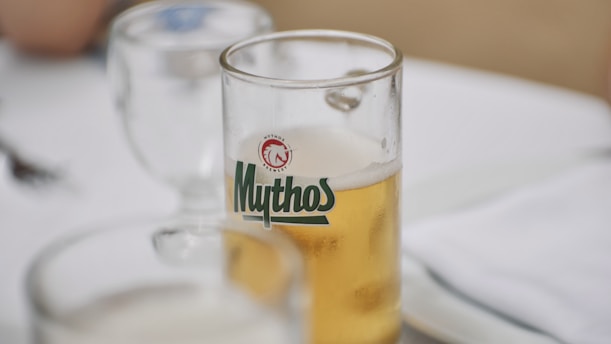 A clear glass filled with golden beer, branded with the logo 'Mythos,' is placed on a white tablecloth. A partially filled wine glass and silverware are also visible on the table.