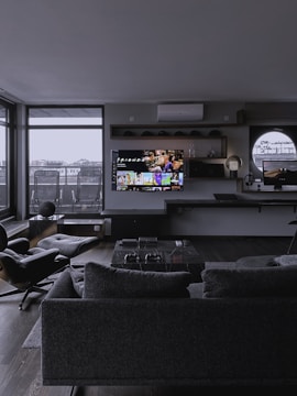 A modern living room with dark, minimalist decor. The area features a large gray sofa, a stylish black lounge chair, and a sleek marble coffee table. A wall-mounted television displays a streaming service interface. Shelves with decorative items and a work desk with a computer monitor are visible. Floor-to-ceiling windows provide a view of an urban balcony with seating.