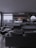 A modern living room with dark, minimalist decor. The area features a large gray sofa, a stylish black lounge chair, and a sleek marble coffee table. A wall-mounted television displays a streaming service interface. Shelves with decorative items and a work desk with a computer monitor are visible. Floor-to-ceiling windows provide a view of an urban balcony with seating.