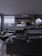 A modern living room with dark, minimalist decor. The area features a large gray sofa, a stylish black lounge chair, and a sleek marble coffee table. A wall-mounted television displays a streaming service interface. Shelves with decorative items and a work desk with a computer monitor are visible. Floor-to-ceiling windows provide a view of an urban balcony with seating.