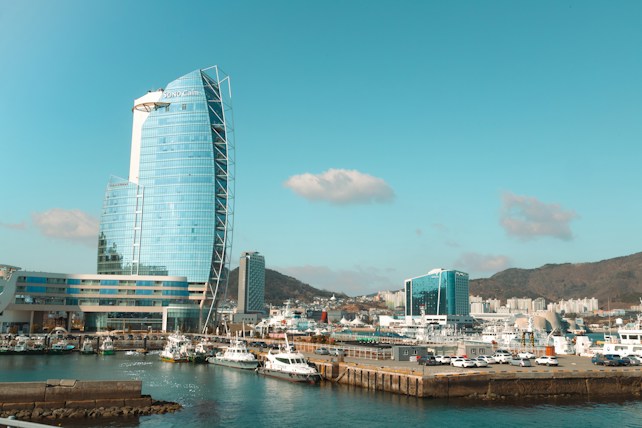 A modern, curved glass skyscraper is situated next to a harbor filled with boats. In the background, there are hills and a cityscape with several buildings. The sky is clear, accentuating the blue color of the skyscraper and the water.