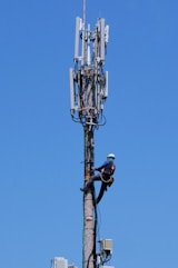 A worker wearing safety gear is climbing a tall communication tower. The tower is equipped with multiple antennas and cables against a clear blue sky.