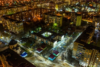 A nighttime aerial view of a city neighborhood features multi-story apartment buildings with numerous lit windows. The buildings are arranged in a grid-like pattern, interspersed with trees and small roads. Streetlights and building lights illuminate the scene, casting a yellowish glow. Few patches of greenery and parked cars are visible along the streets.