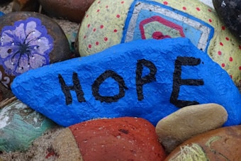 A group of painted rocks, with the focal point being a blue rock with the word 'HOPE' written on it in black letters. Surrounding rocks are decorated with various designs including flowers and abstract patterns.