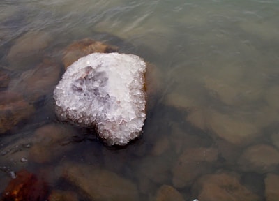 A partially submerged rock is covered with a thick layer of ice, sitting in calm water. The surface of the ice has a rough texture, creating a natural, crystalline appearance.