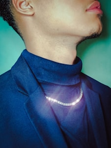 A person's profile is shown wearing a dark blue blazer and a turtleneck, with a sparkling silver chain necklace around the neck. The background is a soft green color.