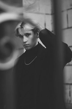 A black-and-white portrait of a young person with light hair, wearing a dark turtleneck and a chain necklace. The background consists of a wall with large tiles, creating a minimalist and moody atmosphere. The person's hand is raised to their head, adding a casual and contemplative element.