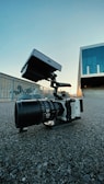 A professional camera with a large lens and external monitor setup is placed on a textured asphalt surface. In the background, there is a modern building with glass panels and a metal railing. The sky is clear with a gradient from blue to light, indicating a sunset.