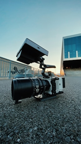 A professional camera with a large lens and external monitor setup is placed on a textured asphalt surface. In the background, there is a modern building with glass panels and a metal railing. The sky is clear with a gradient from blue to light, indicating a sunset.