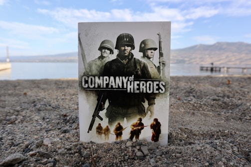 A poster for 'Company of Heroes' is placed upright on a sandy beach with a serene lake and mountains in the background. The poster features three soldiers in military gear and a bold title.