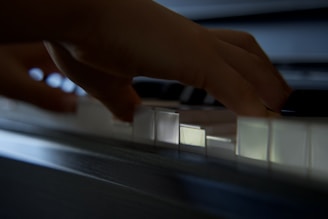 Hands are gracefully playing on the piano keys, casting a soft shadow on them. The image captures the delicate touch and movement of fingers as they press down on the white and black keys. The lighting is subtle, adding to the intimate and focused atmosphere.