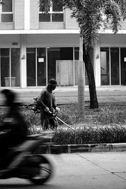 A person wearing a hat and mask uses a string trimmer to cut grass on a sidewalk. A motion-blurred motorcycle passes by in the foreground. In the background, there is a modern building with large windows and a palm tree.
