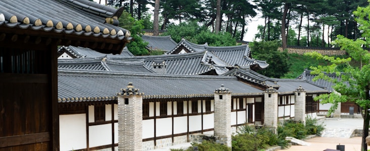 A traditional Korean building featuring tiled roofs and brick columns, set amidst a lush green environment with tall trees in the background.