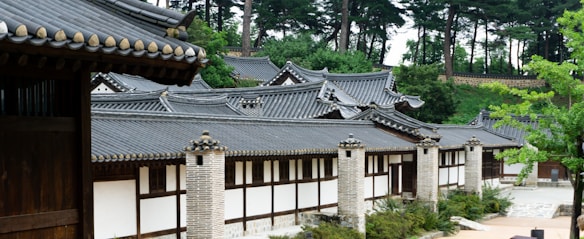 A traditional Korean building featuring tiled roofs and brick columns, set amidst a lush green environment with tall trees in the background.