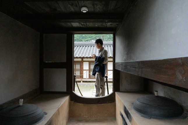 A person stands outside, framed by the interior of a traditional building with wooden beams and large pots or stoves visible on each side. The person appears to be holding a jacket and looking at a smartphone, with a courtyard and tiled roofs in the background, suggesting a historical or cultural setting.