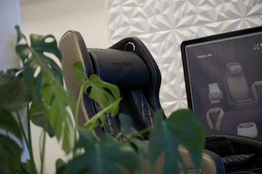 A black and beige massage chair is positioned next to lush green leaves of a monstera plant. The backdrop features a patterned white wall, and a monitor displays images of various massage devices.