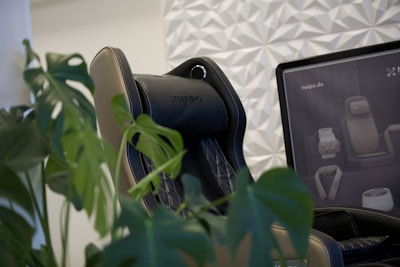 A black and beige massage chair is positioned next to lush green leaves of a monstera plant. The backdrop features a patterned white wall, and a monitor displays images of various massage devices.