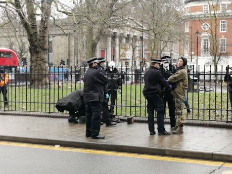 Several police officers interact with a person wearing camouflage clothing next to a metal fence on a city street. A London double-decker bus and old buildings are visible in the background. The scene suggests a potentially tense situation.