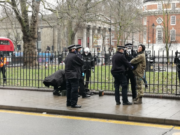 Several police officers interact with a person wearing camouflage clothing next to a metal fence on a city street. A London double-decker bus and old buildings are visible in the background. The scene suggests a potentially tense situation.