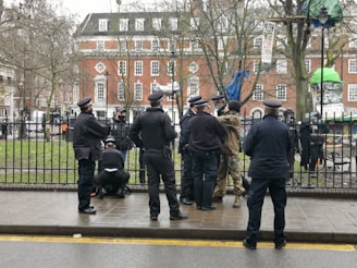 A group of six uniformed officers stand on a wet street near a fenced park. They appear to be having a discussion, and one officer is kneeling. In the background, an individual in camouflage clothing is also engaged with the officers. Various signs and banners are visible in the park area, hanging from trees near colorful tents. Leafless trees and a large brick building can be seen in the background.