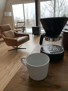 A snowy landscape viewed from inside a warmly lit room, with a steaming mug on the windowsill and a woolen throw on the armchair.