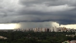 Dark storm clouds loom over a city skyline, with a prominent funnel cloud extending towards the ground. The city is surrounded by dense green foliage.