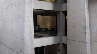A modern building with visible concrete surfaces and geometric design. The interior is partially visible through large glass windows, revealing a seating area with chairs. The walls inside feature vertical lines and muted lighting.