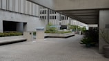 A modern urban courtyard with concrete architecture, featuring low walls and benches. The space is open with some greenery, including potted plants and shrubs. The pavement has a geometric pattern, and the area is partially covered by a concrete overhang. In the background, there are large office buildings with multiple stories.