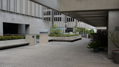 A modern urban courtyard with concrete architecture, featuring low walls and benches. The space is open with some greenery, including potted plants and shrubs. The pavement has a geometric pattern, and the area is partially covered by a concrete overhang. In the background, there are large office buildings with multiple stories.