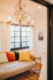 A cozy living space featuring a beige tufted sofa adorned with colorful cushions in shades of blue, orange, and yellow. A decorative, ornate chandelier hangs from the ceiling, casting warm light over the room. Light filters in through large windows with white curtains framing them. A small round side table stands next to the sofa, and a framed picture hangs on the wall.