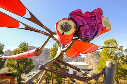 A large, colorful sculpture resembling an insect with red and pink hues, set against a backdrop of mountains and greenery. The sculpture features prominent red wings and a purple head, with intricate metal framework.