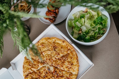 A cheese pizza in a cardboard box is placed on a surface surrounded by other dishes. A fresh green salad is in a bowl to the side, and a plate with skewered grilled food is also present. Greenery is partially visible at the top of the image, adding a touch of nature.