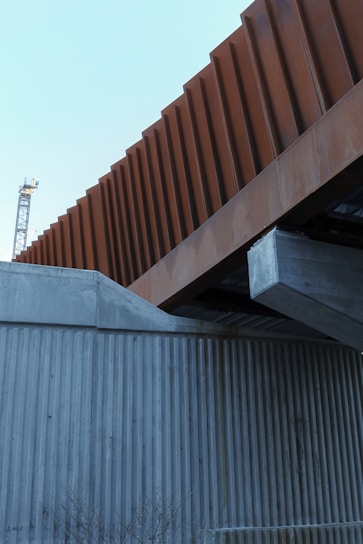A modern architectural structure featuring intersecting elements made of concrete and weathered steel. The foreground includes a concrete wall with vertical grooves, while above it, a prominent steel beam with a rust-colored facade contrasts against the sky. In the background, part of a crane is visible, indicating a construction area.
