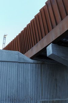 A modern architectural structure featuring intersecting elements made of concrete and weathered steel. The foreground includes a concrete wall with vertical grooves, while above it, a prominent steel beam with a rust-colored facade contrasts against the sky. In the background, part of a crane is visible, indicating a construction area.