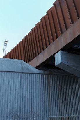 A modern architectural structure featuring intersecting elements made of concrete and weathered steel. The foreground includes a concrete wall with vertical grooves, while above it, a prominent steel beam with a rust-colored facade contrasts against the sky. In the background, part of a crane is visible, indicating a construction area.