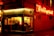 An inviting corner café emits a warm, welcoming glow from its neon sign and interior lights. People are seated inside, enjoying conversation, and the scene suggests a cozy, bustling atmosphere typical of a popular café.