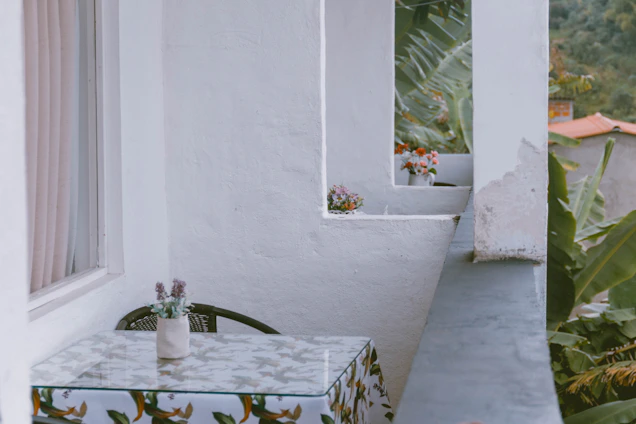 A sunlit balcony featuring a compact rattan bistro set with a small metal table and a wall-mounted plant shelf adorned with greenery.
