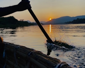 A paddle is being used to row a boat on a calm river during sunset. The sunlight reflects off the water, creating a serene and peaceful ambiance. The river is surrounded by greenery and hills under the warm glow of the setting sun.