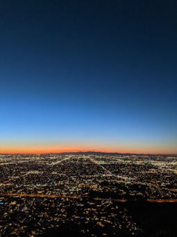 A breathtaking panoramic view of a cityscape at dusk, with a vast array of sparkling lights sprawling across the landscape. The sky displays a gradient of colors from deep blue at the top to a warm orange near the horizon.