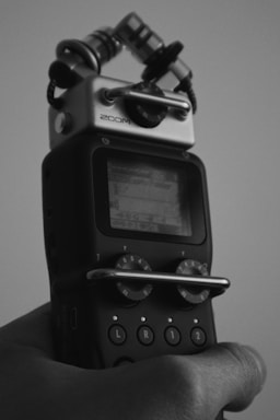 A black and white image features a handheld audio recording device with visible dials and buttons. The screen displays some digital information, though it's not clearly readable. The device is gripped by a hand.