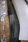 A modern architectural interior with large, curved glass panels and smooth, flowing walls. The design showcases a blend of metal, glass, and stone elements with natural light filtering through expansive windows. The space appears open and airy, with a focus on geometric and organic forms.