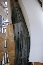 A modern architectural interior with large, curved glass panels and smooth, flowing walls. The design showcases a blend of metal, glass, and stone elements with natural light filtering through expansive windows. The space appears open and airy, with a focus on geometric and organic forms.