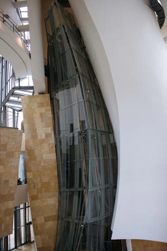 A modern architectural interior with large, curved glass panels and smooth, flowing walls. The design showcases a blend of metal, glass, and stone elements with natural light filtering through expansive windows. The space appears open and airy, with a focus on geometric and organic forms.
